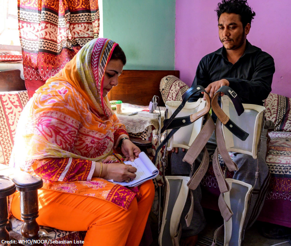 A fieldworker writing on a paper notepad sitting next to a disabled person. Cover Image