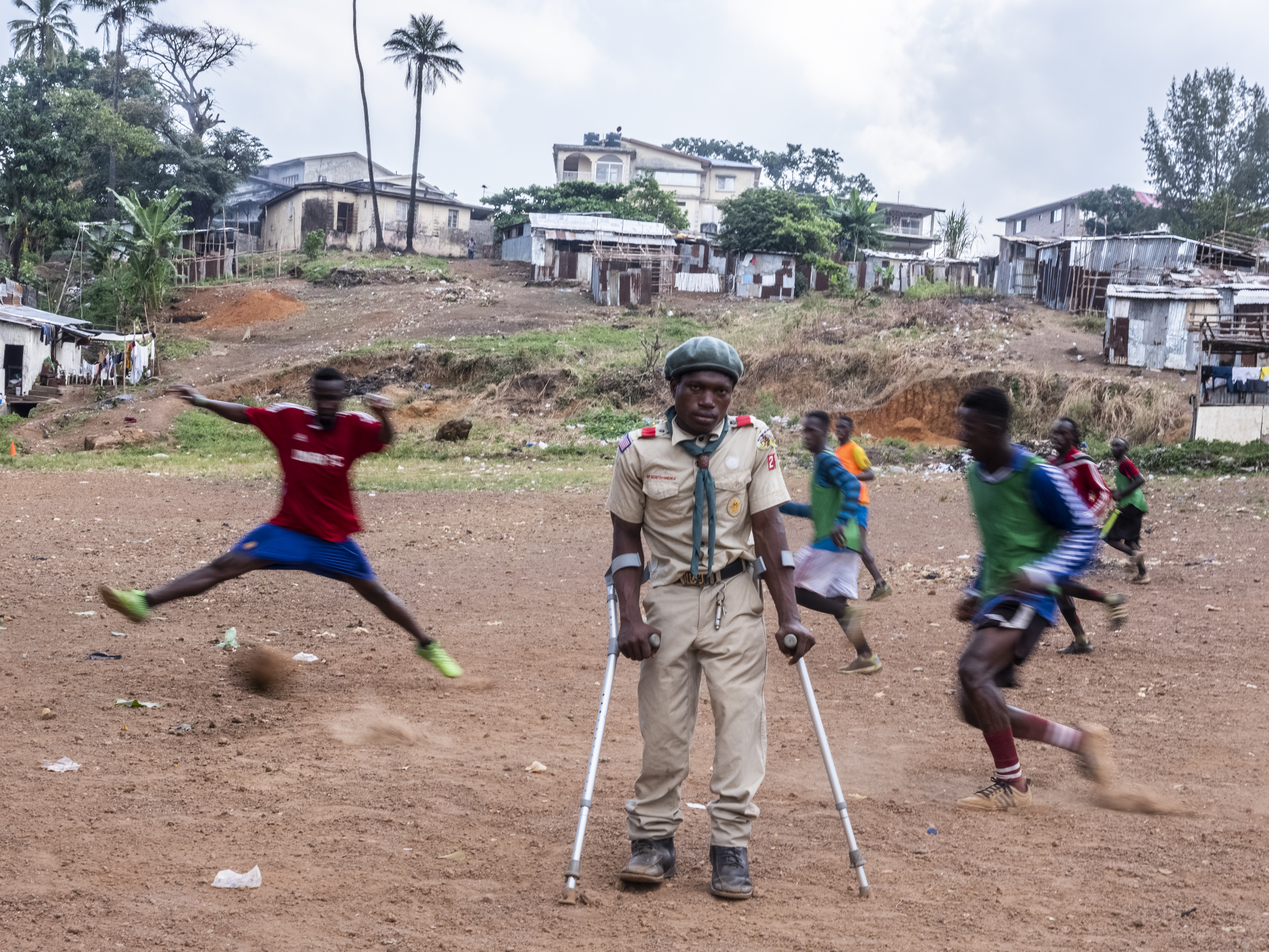 Man using an assistive tech device in the centre of playground with young people playing football behind him Cover Image