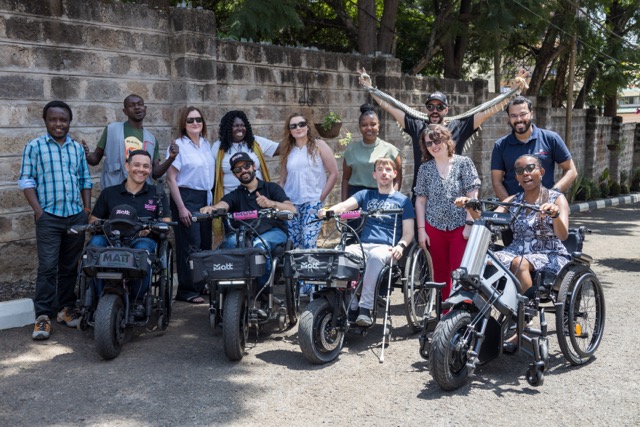 A group of people pose outdoors on a paved street beside a stone wall, with several individuals seated on electric wheelchairs and handcycles labeled “MATT,” while others stand behind them. Trees line the background. Cover Image