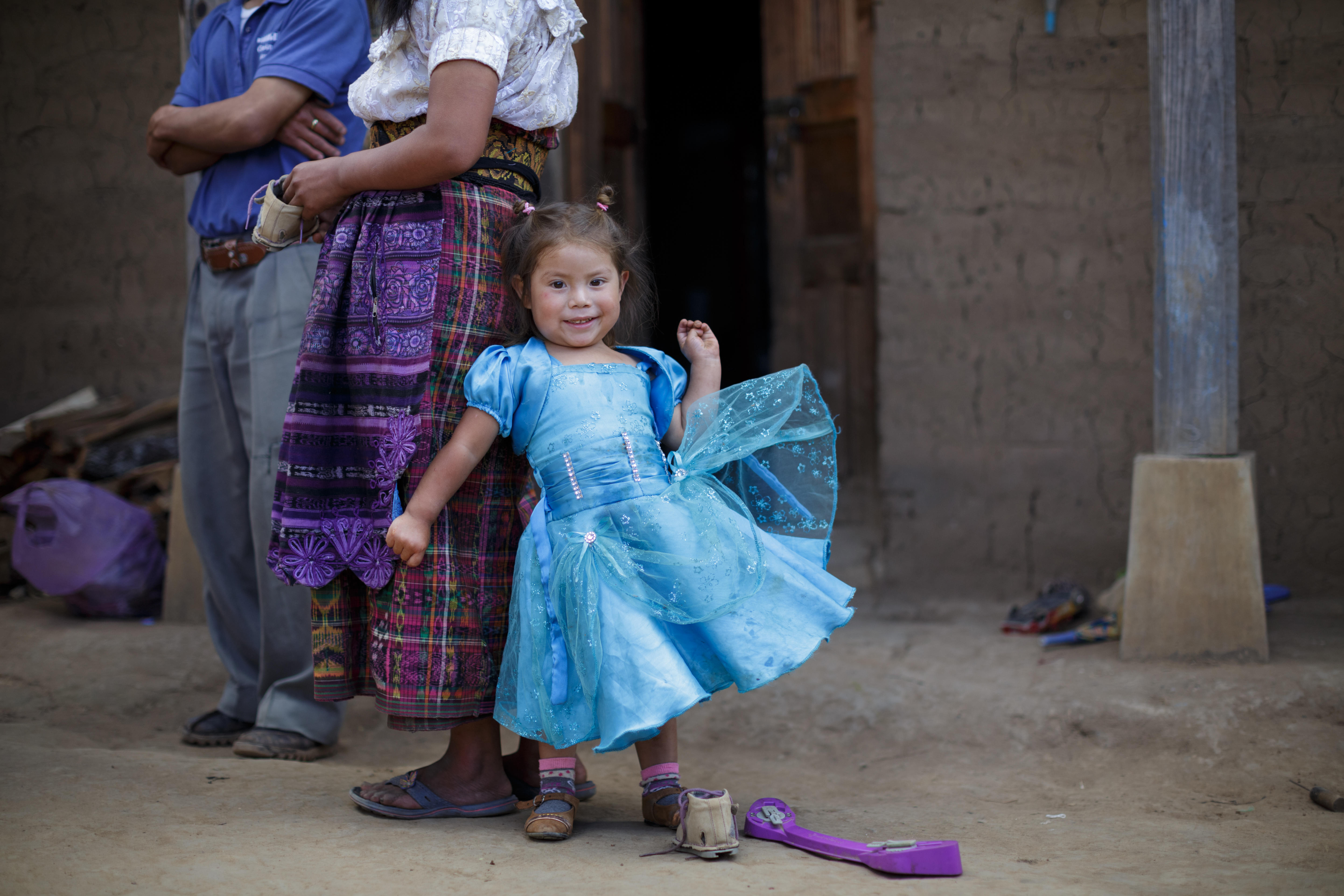 A young girl in blue dress standing next to a woman smiling, Miracle Feet brace and shoe on the floor next to her