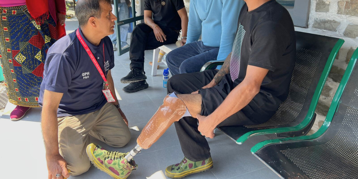 A person sitting on a bench being fitted with a liner and prosthetic leg. They are talking to a clinician and have other participants and clinicians nearby.