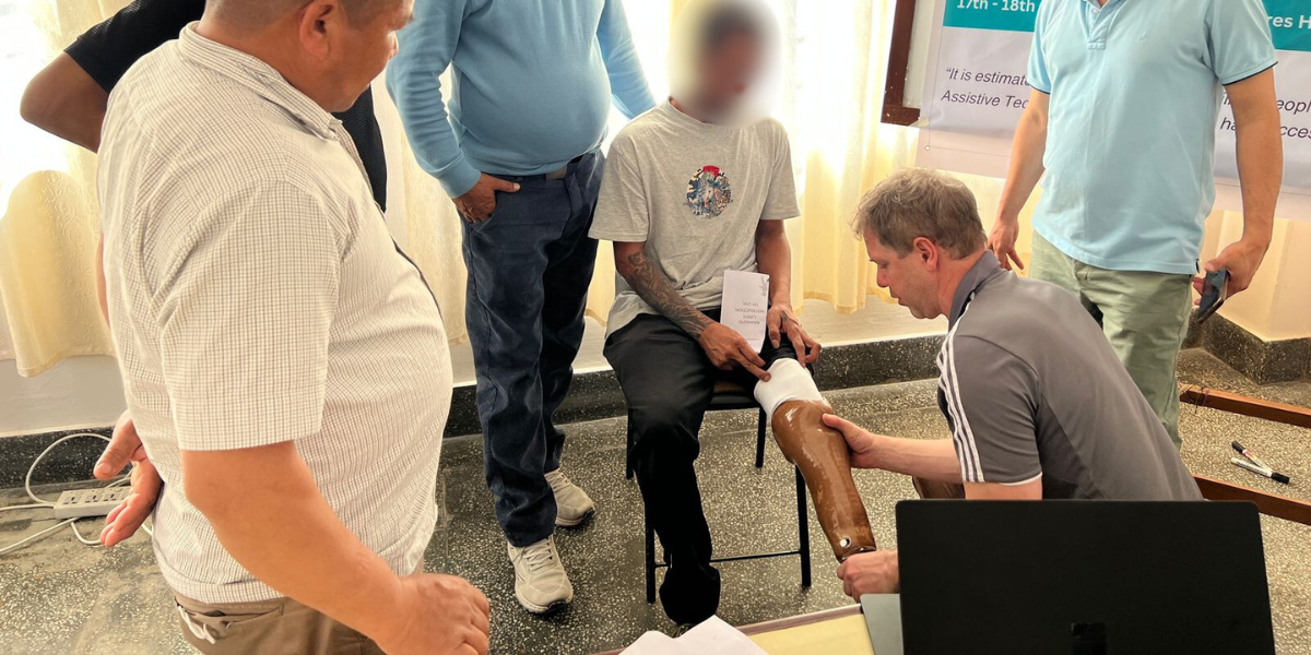 A person is being fitted with a prosthetic leg while they sit in a chair this is being carried out by commission while other clinicians watch