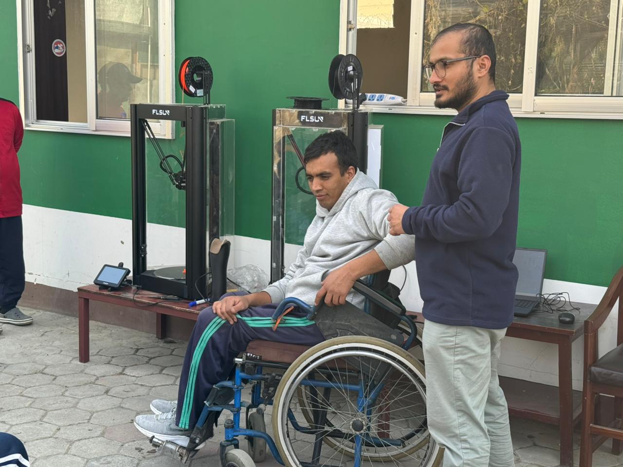 A person in a wheelchair sits near two 3D printers outdoors while another person stands beside them.