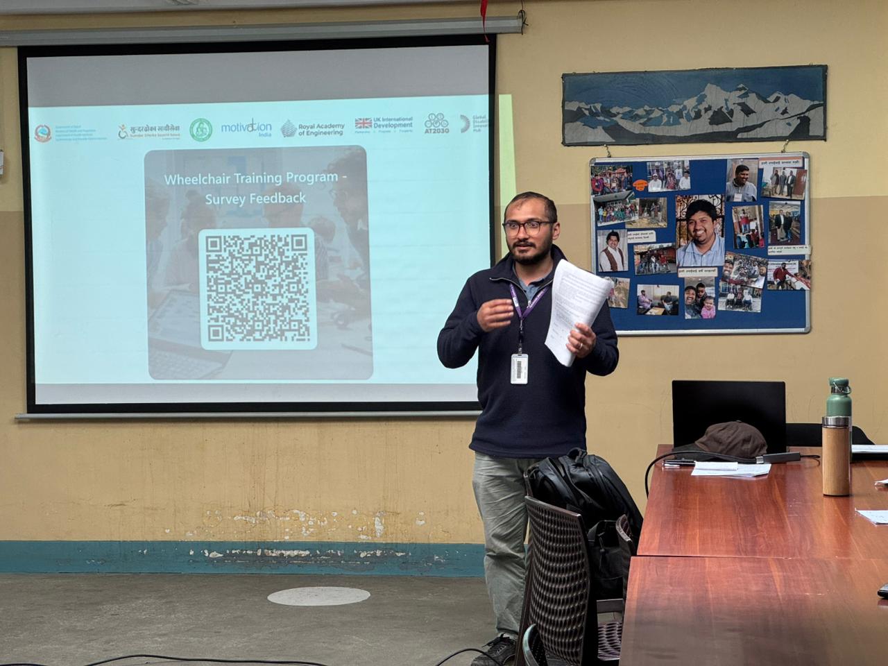 Ram stands presenting in front of a projected slide titled “Wheelchair Training Program – Survey Feedback,” holding papers.
