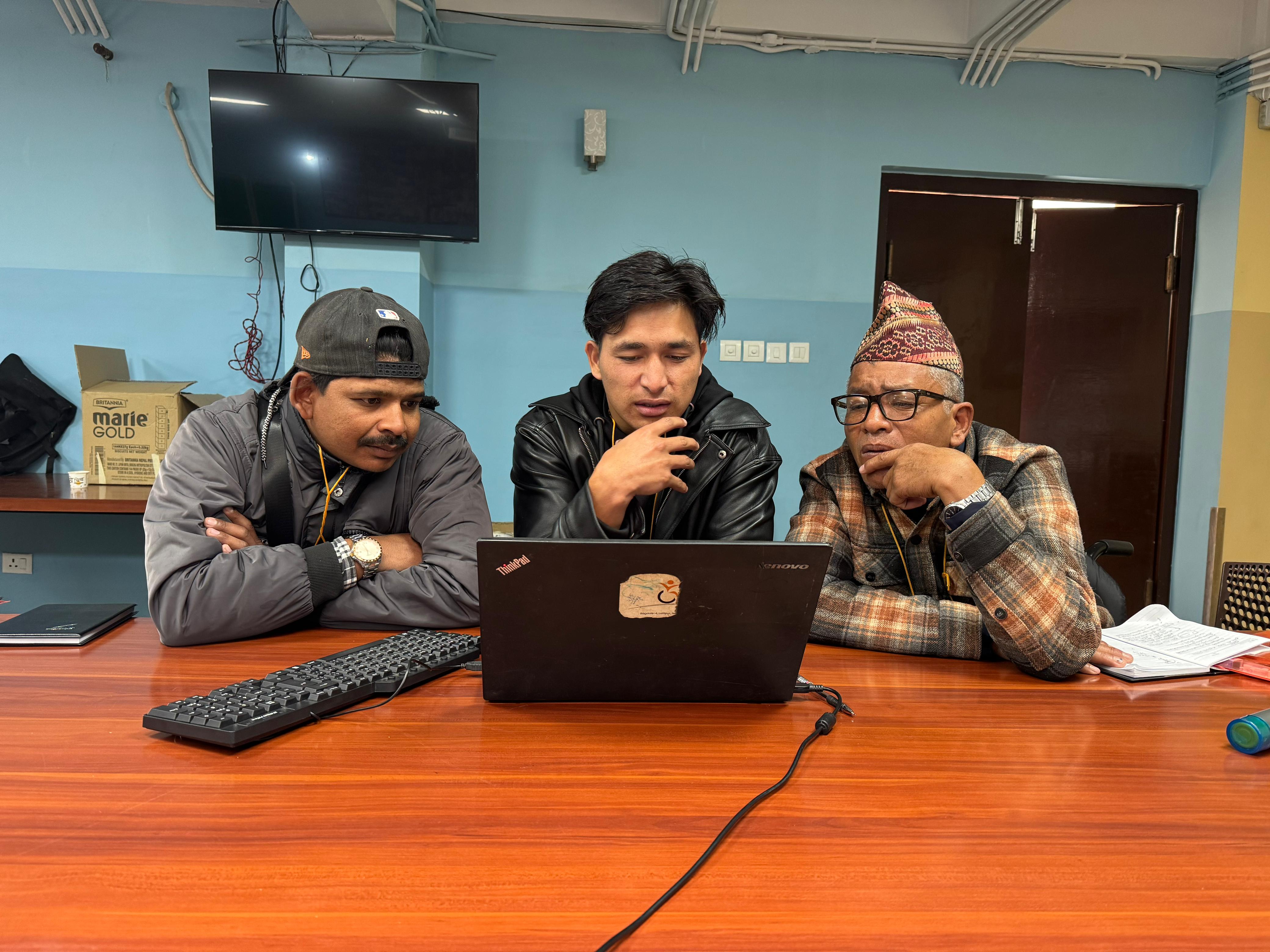 Three people sit at a table working together on a laptop, with a keyboard in front of them in a meeting room.