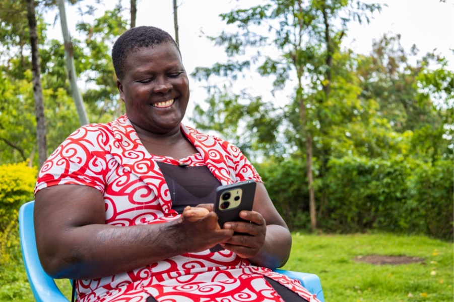 Carolyne sat outside smiling. They have an impairment on their left hand and are using a mobile phone. 