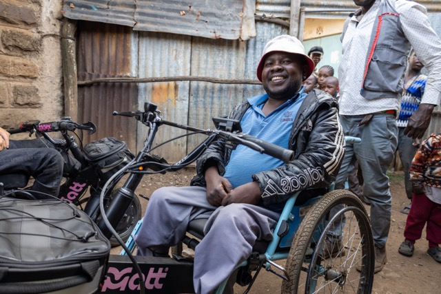 A person sits on a hand‑powered wheelchair next to an electric handcycle labeled &ldquo;MATT,&rdquo; on a narrow street with corrugated metal walls and several people standing in the background.