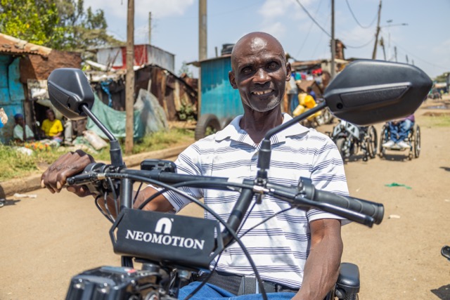 A person sits on an electric handcycle labeled &ldquo;NEOMOTION&rdquo; on a dirt road, with multiple handcycles lined up behind them and small roadside structures in the background.