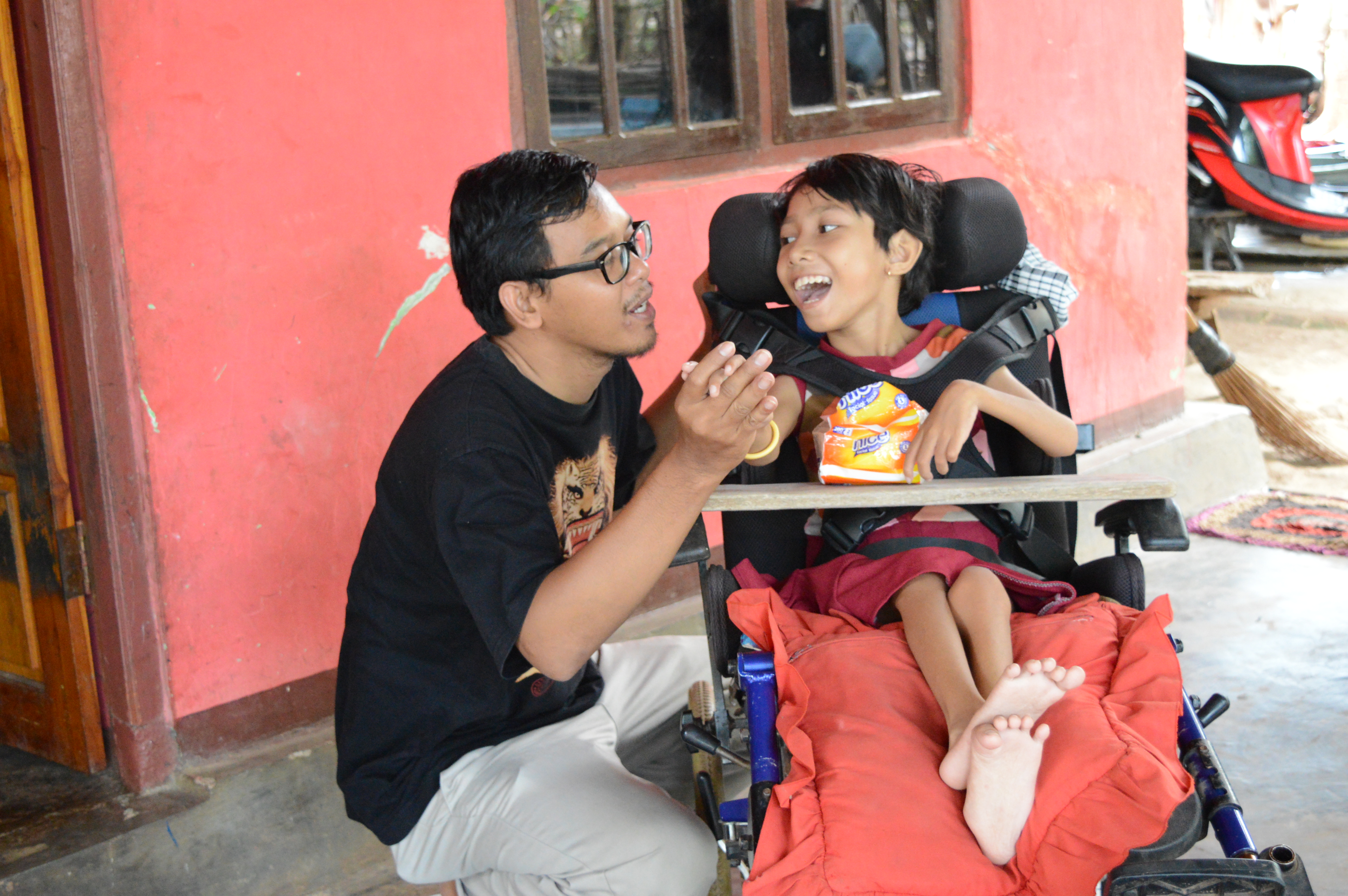 Eka is knelt down next to a younger child who is using a wheelchair. The child is looking at Eka and smiling