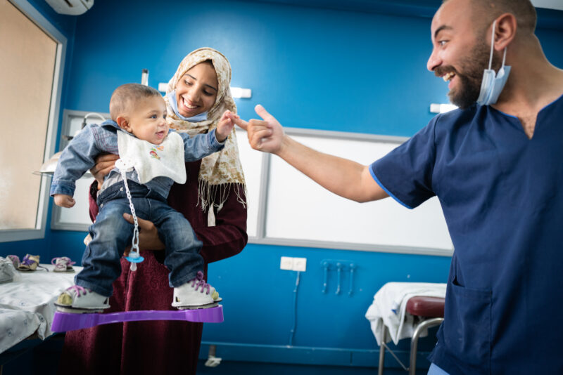 A child is wearing the MiracleFeet brace. The brace is attached to his shoes and keeps his feet in a turned out position. The child is being held by his mother and smiling at a clinician, grabbing his hand Cover Image