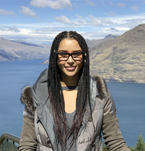 Naomi Thompson wearing glasses and a grey jacket, standing on a hilltop with a lake and mountains in the background